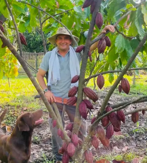 A grower in a sun hat smiling among cacao trees heavy with red pods, a dog at his side
